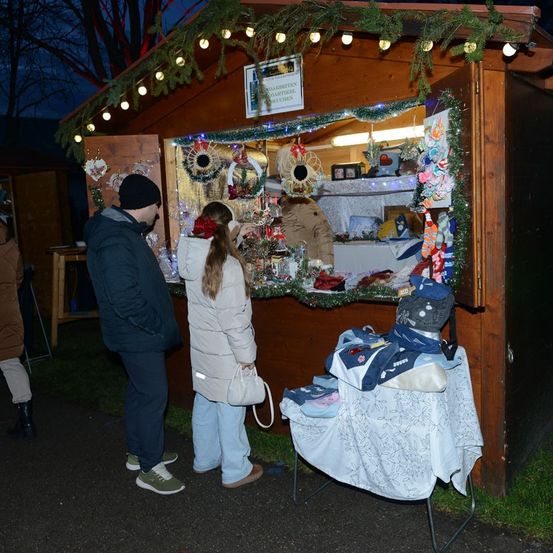 Ein Paar steht vor einem festlich geschmückten Stand in einem Park, verziert mit Girlanden und Lichtern. Im Inneren sind verschiedene Winterartikel ausgestellt.