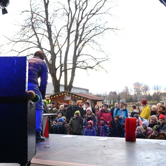 Eine Menschenmenge steht in einem Freiluftbereich und beobachtet einen Mann in Blau auf der Bühne. Die Bühne hat eine blaue Box und einen roten Gegenstand. Bäume und ein Gebäude sind im Hintergrund.