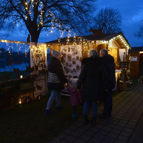 Eine Familie steht vor einem dekorierten Weihnachtsstand mit Lichterketten. Der Stand hat einen See und Bäume im Hintergrund.
