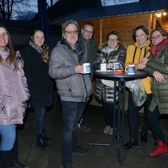 Eine Gruppe von Menschen versammelt sich bei Dämmerung um einen Tisch in einem Schuppen und hält Tassen. Sie lächeln und scheinen ein Gespräch zu führen.