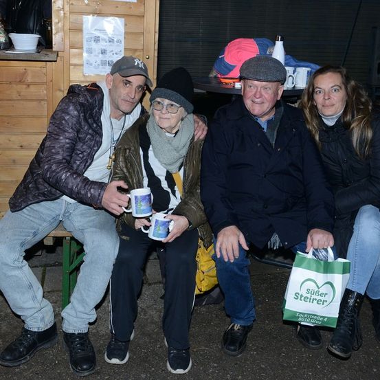 Vier Menschen sitzen zusammen und lächeln. Zwei Männer tragen Jacken und halten Tassen, einer hat eine grüne Tasche. Eine ältere Frau trägt eine Brille und einen Schal. Hinter ihnen steht eine Holzstruktur mit einem Becher und einem Papier.