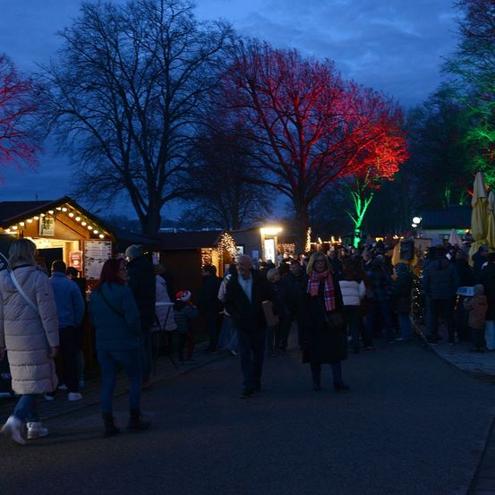 Eine Menschenmenge geht bei Nacht eine Straße entlang, gesäumt von beleuchteten Bäumen und Weihnachtsständen.