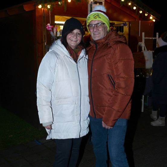 Ein Mann und eine Frau stehen zusammen vor einem Weihnachtsstand und lächeln. Die Frau trägt einen weißen flauschigen Mantel und der Mann einen braunen flauschigen Mantel und eine Mütze mit einem Grinch-Design.