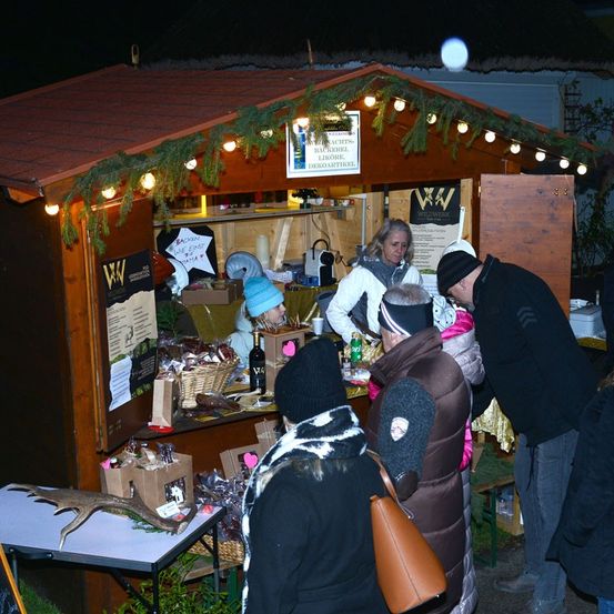 Menschen versammeln sich um einen mit Weihnachtslichtern geschmückten Holzkiosk. Der Kiosk ist mit Artikeln wie Likör, Süßigkeiten und Weinflaschen bestückt. Ein Hirschkopf ist auf einem nahegelegenen Tisch ausgestellt.