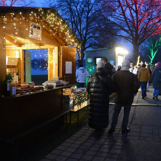 Ein Weihnachtsmarkt mit einem kleinen, mit Lichtern geschmückten Laden, an dem Menschen bei Dämmerung vorbeigehen.