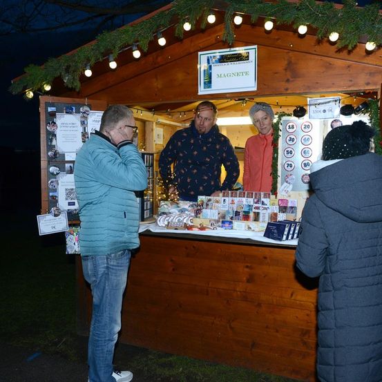 Ein hölzerner Stand bei Nacht mit Weihnachtsdekorationen zeigt zwei Männer und eine Frau. Ein Mann telefoniert, während ein anderer Mann und eine Frau hinter dem Tresen stehen.