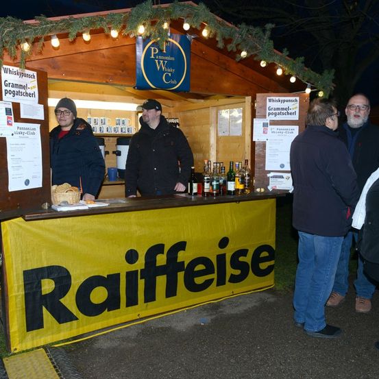Eine Gruppe von Menschen versammelt sich um einen Stand, der Whisky verkauft. Er ist mit festlichen Lichtern und einem Schild, das Raiffeisen steht, dekoriert.