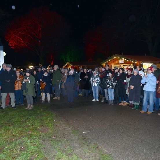Eine Menschenmenge steht bei Nacht im Freien, beleuchtet von roten Lichtern auf Bäumen. Sie scheinen an einer Veranstaltung teilzunehmen, einige halten Tassen. Das Gebiet ist von Ständen mit Weihnachtslichtern umgeben.