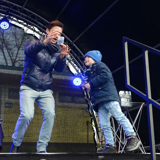 Ein Zauberer führt auf der Bühne mit einem kleinen Jungen vor. Der Zauberer hält eine Karte in der Hand, während der Junge aufmerksam zusieht. Die Bühne ist mit blauen Lichtern beleuchtet.