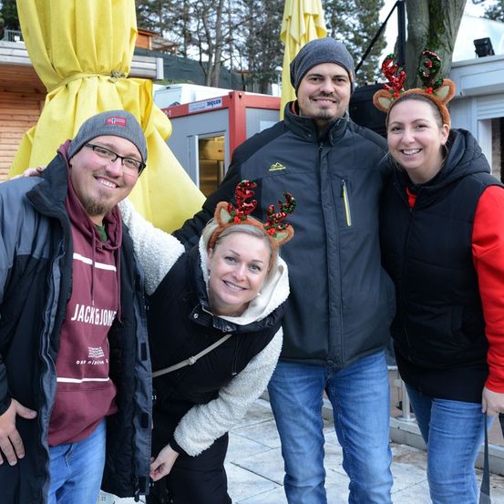 Vier Menschen in Winterkleidung lächeln und posieren für ein Foto. Zwei Männer und zwei Frauen, alle mit Weihnachtskopfbändern, stehen eng zusammen vor einem Holzgebäude.