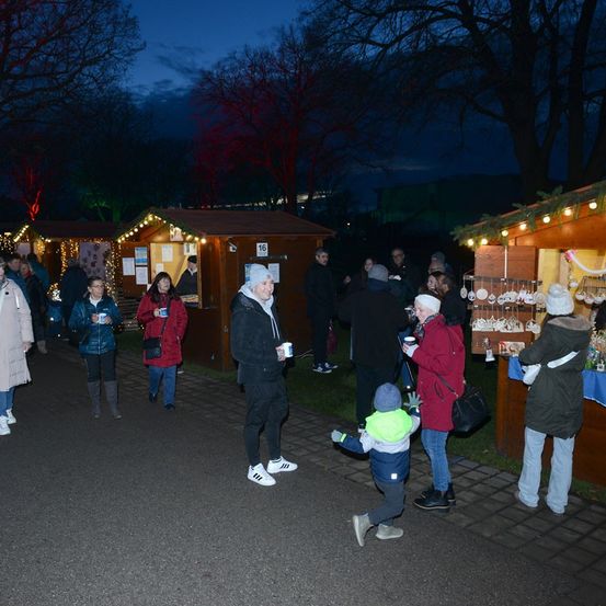 Bei Dämmerung versammeln sich Menschen um beleuchtete Stände in einem Park, einige halten Tassen, andere schauen sich die Artikel an. Bäume und Gebäude bilden den Hintergrund.