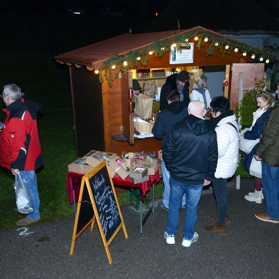 Ein kleiner Holzstand, dekoriert mit Weihnachtslichtern, ist nachts geöffnet. Einige Leute sind um den Stand versammelt und kaufen etwas. Der Stand hat ein Schild und eine Tafel mit Schrift.