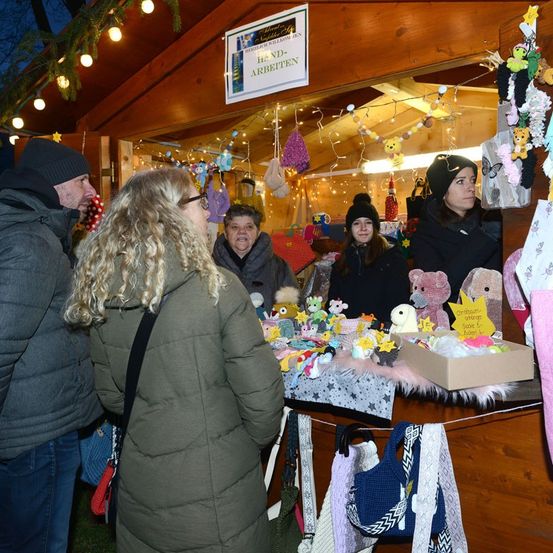 Eine Gruppe von Menschen steht an einem festlich dekorierten Marktstand mit Plüschtieren und Winterkleidung. Sie schauen sich die Artikel an und der Stand ist mit bunten Lichtern beleuchtet.
