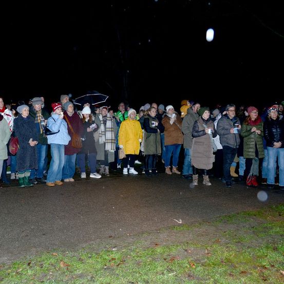 Eine Menschenmenge, in Winterkleidung gekleidet, steht in einer Reihe an einem dunklen Abend und hält Tassen und einige mit Regenschirmen.