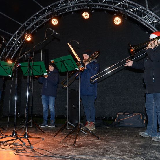 Eine musikalische Aufführung findet nachts statt. Drei Personen spielen Posaunen und eine Trompete. Sie stehen auf einer Bühne mit Notenständern und Mikrofonen. Scheinwerfer sind über ihnen.