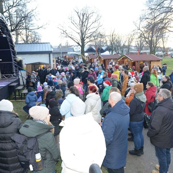 Eine Menschenmenge versammelt sich im Freien in der Nähe einer Bühne mit festlichen Lichtern. Viele Menschen tragen Winterkleidung, Mützen und Brillen. Einige haben Rucksäcke und halten Tassen. Kleine Hütten mit Lichtern sind im Hintergrund zu sehen.