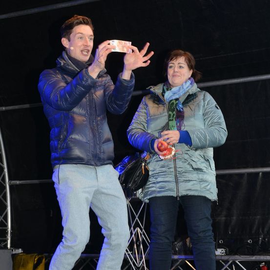 Ein Mann und eine Frau auf der Bühne bei Nacht, der Mann hält eine 10-Euro-Banknote und spricht, die Frau hält einen Apfel und lächelt.