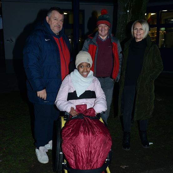 Vier Menschen stehen bei Nacht draußen. Eine junge Frau im Rollstuhl ist in der Mitte. Hinter ihr stehen zwei Männer und eine Frau, alle lächeln.