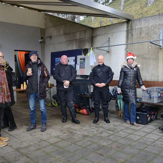 Fünf Menschen stehen in einem Raum. Drei von ihnen tragen schwarze Anzüge. Einer von ihnen hält einen Becher. Die Frau rechts trägt einen Weihnachtsmannhut. Es gibt einige Lagerboxen und einen Tisch mit einigen Gegenständen darauf.