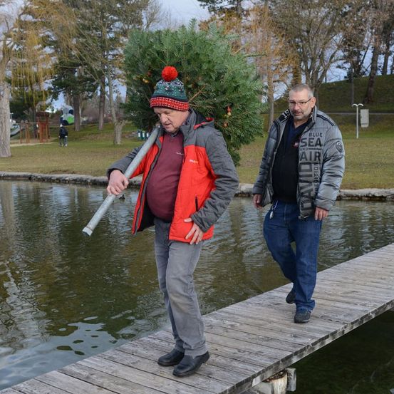 Zwei Männer gehen auf einer Holzbrücke. Einer trägt einen Weihnachtsbaum, während der andere eine Brille und eine Jacke trägt. Der Hintergrund zeigt einen Teich, Bäume und einen Spielplatz.