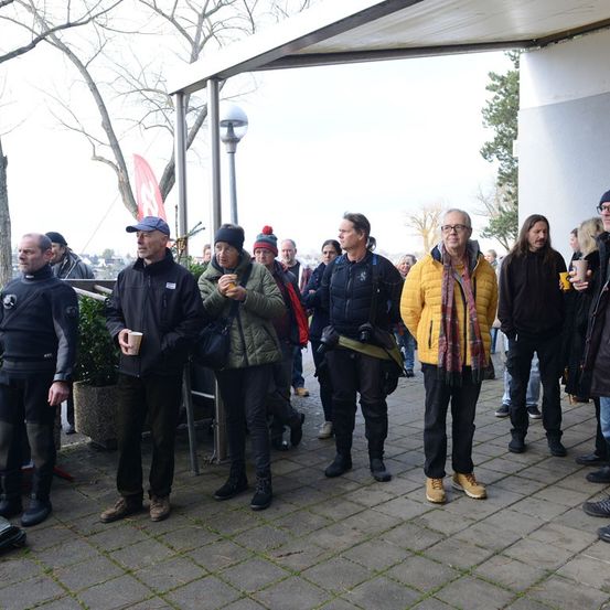 Eine Gruppe von Menschen, eingemummelt in Winterkleidung, steht auf einem gepflasterten Weg unter einem Dach. Einige halten Tassen. Eine Person trägt eine blaue Kappe. Im Hintergrund befinden sich Bäume und ein Gebäude.