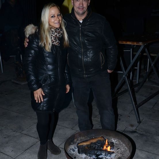 Ein Mann und eine Frau stehen eng beieinander vor einem Feuerkorb und lächeln für ein Foto. Der Mann hat seinen Arm um die Schulter der Frau gelegt. Dahinter steht ein Tisch mit einem Stuhl.