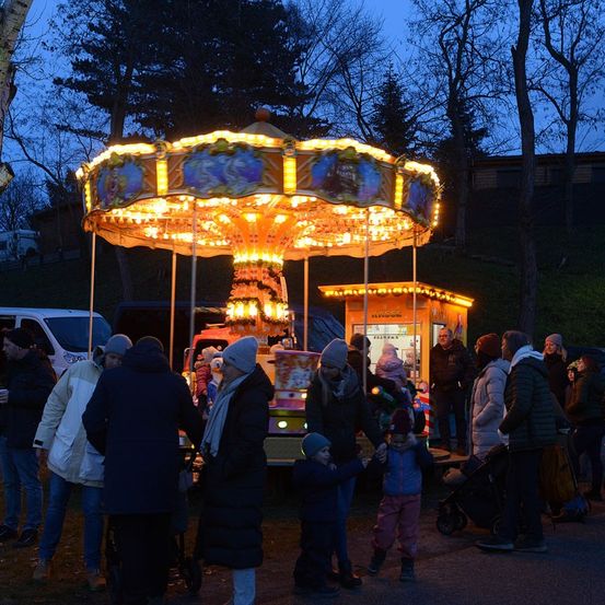 Ein Karussell mit bunten Lichtern ist nachts beleuchtet. Menschen, einschließlich Kinder, versammeln sich darum. Einige halten Händchen, während andere in der Nähe des Karussells und der Stände stehen.