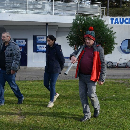 Drei Personen gehen auf einem Rasenbereich in der Nähe eines Gebäudes. Ein Mann trägt einen geschmückten Tannenbaum. Eine Frau in einer Jacke und Jeans folgt ihm. Hinter ihnen ist ein Gebäude mit einem Schild, das TAUCH liest.