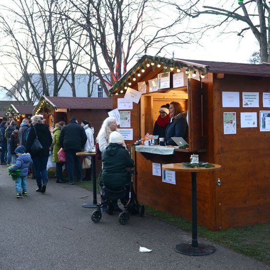 Eine Schlange von Menschen wartet darauf, eine kleine Holzhütte mit Weihnachtslichtern zu betreten. Eine Frau und ein Kind sitzen an der Theke.
