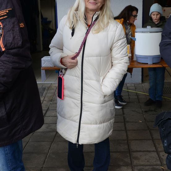 Eine Frau mit blonden Haaren, die eine weiße gepolsterte Jacke trägt, steht mit den Händen in den Taschen. Hinter ihr hält eine andere Frau einen weißen Reiskocher.