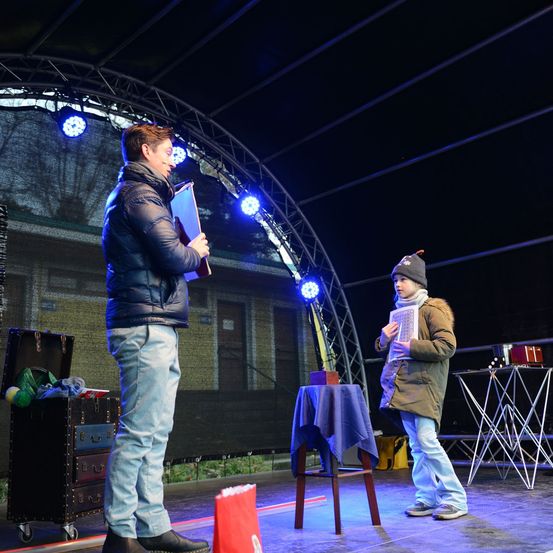 Ein Mann und ein Kind stehen auf der Bühne unter blauen Lichtern. Der Mann hält eine Mappe und das Kind ein Buch. Es gibt Kisten, einen Tisch und Stühle auf der Bühne.