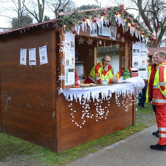Ein Mann in einer reflektierenden Weste sitzt lächelnd an einem kleinen Holzkiosk. Ein zweiter Mann steht in der Nähe, und beide sind draußen an einem sonnigen Tag.