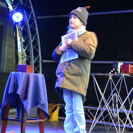 Ein junges Mädchen steht auf der Bühne, hält ein Buch in der Hand, trägt einen Hut, einen Schal und einen Mantel, mit einem blauen Tischtuch und einer Metallstruktur hinter ihr.