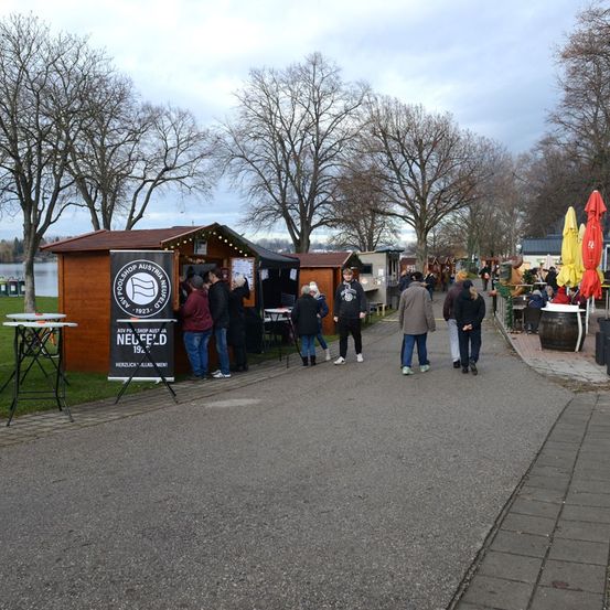 Ein Weg mit mehreren kleinen Holzhütten ist zu sehen, an dem Menschen vorbeigehen. Es gibt blattlose Bäume und geschlossene Regenschirme.
