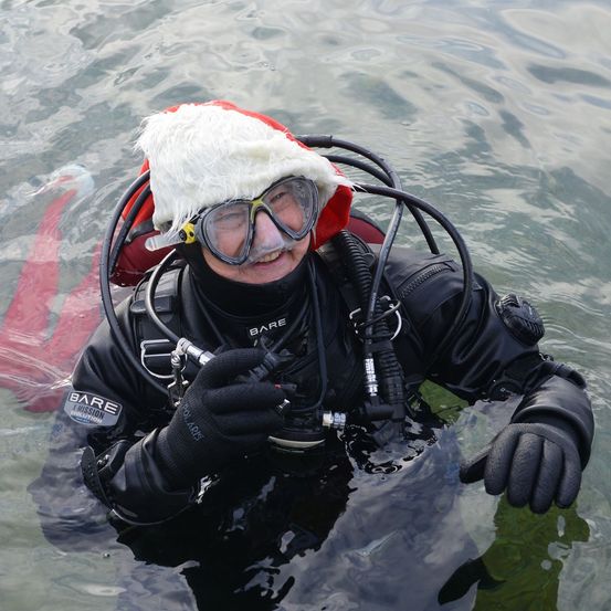 Eine Person in Tauchausrüstung, die einen Weihnachtsmannhut trägt, lächelt unter Wasser. Der Taucher hat eine Maske, einen Atemregler und Handschuhe. Der Anzug hat die Marke 'BARE'.