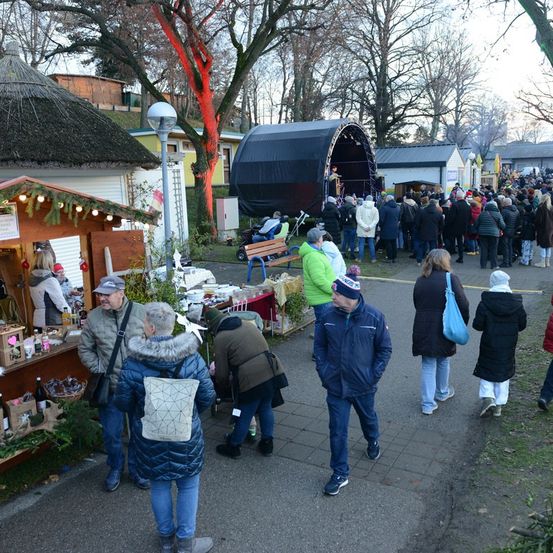 Eine Menschenmenge geht an Essensständen und einer Bühne in einem Park vorbei. Einige tragen Winterkleidung, andere haben Handtaschen. Bäume und ein Gebäude sind im Hintergrund.