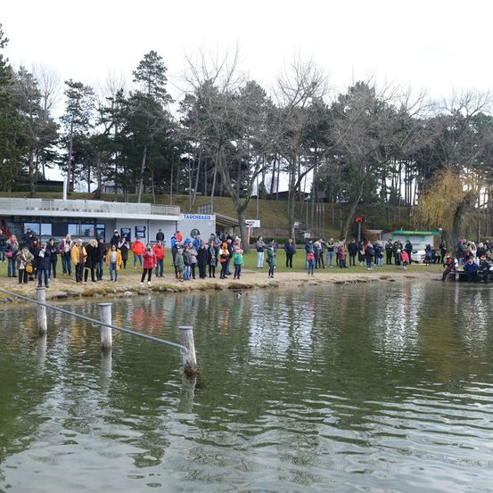 Eine Menschenmenge steht am Ufer eines Sees, einige tragen Mützen und Schuhe. Ein Gebäude mit einem blauen Schild, das 'Tauchbasis' besagt, ist sichtbar. Das Gebiet ist von Bäumen und einem grasbewachsenen Hügel umgeben.