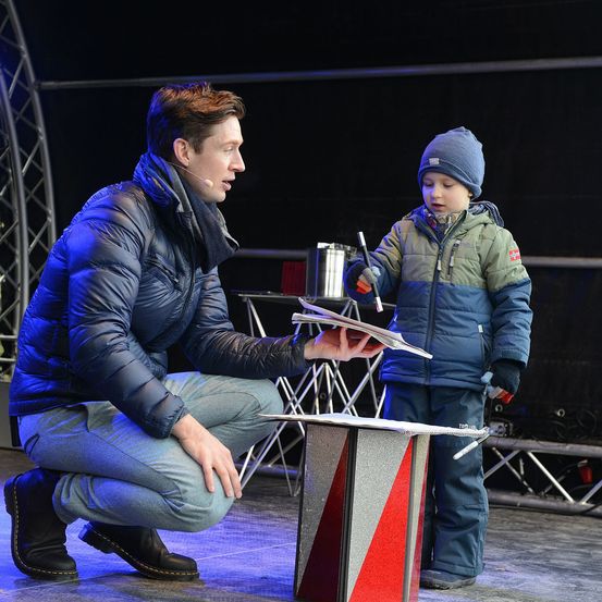 Ein Mann mit Mikrofon kniet und zeigt etwas einem Kind. Das Kind steht neben einer kleinen rot-weiß gestreiften Box.