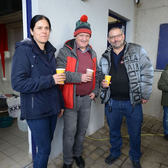 Drei Menschen stehen eng beieinander, lächeln und halten gelbe Becher in der Hand. Der Mann in der Mitte trägt einen roten und grauen Mantel. Der Mann rechts trägt einen grauen Mantel mit dem Wort 'DAVID' darauf. Die Frau links trägt einen blauen Mantel. Sie scheinen für ein Foto zu posieren.