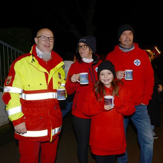 Vier Menschen stehen nachts zusammen, alle in roten Jacken. Zwei Erwachsene sind links und rechts, eine junge Frau in der Mitte. Sie alle halten Tassen.