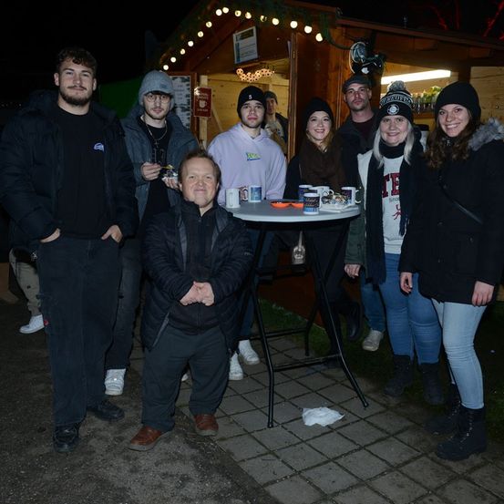 Eine Gruppe von Menschen steht nachts zusammen und lächelt, mit Tassen auf einem Tisch vor ihnen. Dahinter steht ein kleines Häuschen mit Weihnachtslichtern.