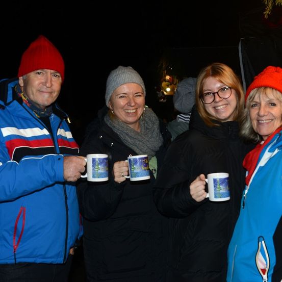 Vier Erwachsene, gekleidet in Winterkleidung, lächeln für ein Foto. Sie halten Tassen mit Weihnachtsdesigns in den Händen. Der Hintergrund ist dunkel, möglicherweise nachts.