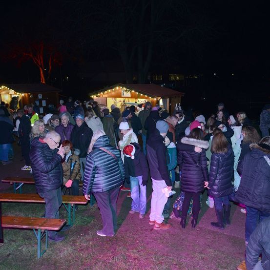 Eine Menschenmenge versammelt sich am Abend draußen, viele in Winterkleidung. Im Hintergrund befinden sich Stände und ein Weihnachtsbaum.