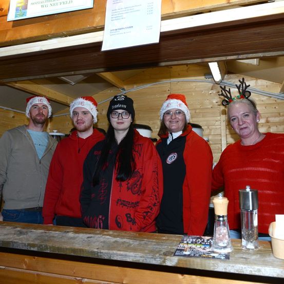 Fünf Erwachsene stehen hinter einem Tresen in roten Jacken und Weihnachtsmützen. Das Schild hinter ihnen lautet 'SOLZAUTHERA NEUFELD.' Auf dem Tresen befinden sich Tassen, Salz und Pfefferstreuer.
