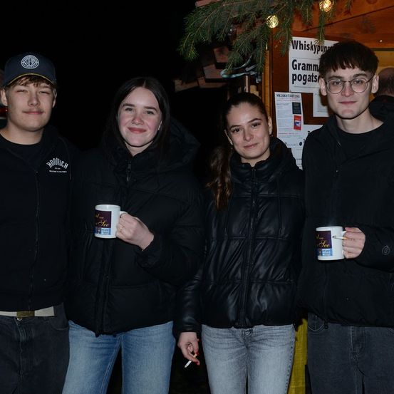 Vier Personen stehen in einer nächtlichen Umgebung zusammen, alle tragen schwarze Jacken. Jeder von ihnen hält eine weiße Tasse mit einem Logo und dem Wort 'See' darauf. Einer von ihnen raucht eine Zigarette. Hinter ihnen steht eine Holzkonstruktion mit einem Schild, auf dem 'Whiskypuns Gramm pogats' steht.