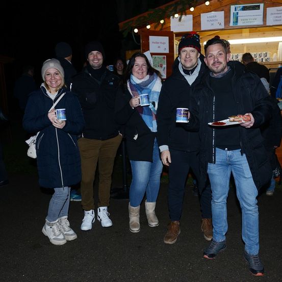 Fünf Menschen in Winterkleidung stehen nebeneinander, halten Tassen und lächeln für die Kamera. Hinter ihnen ist eine Holzstruktur mit Schildern zu sehen.