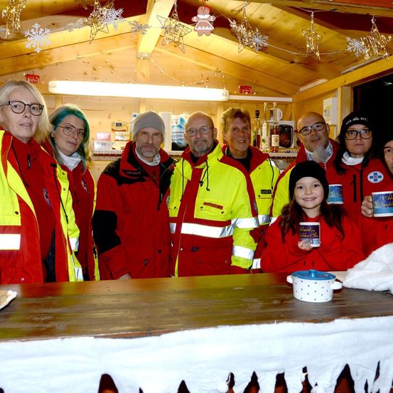 Eine Gruppe von Erwachsenen und einem Kind in roten Jacken steht in einem festlich geschmückten Raum mit einem Holztisch vor ihnen. Sie lächeln und posieren für ein Foto.