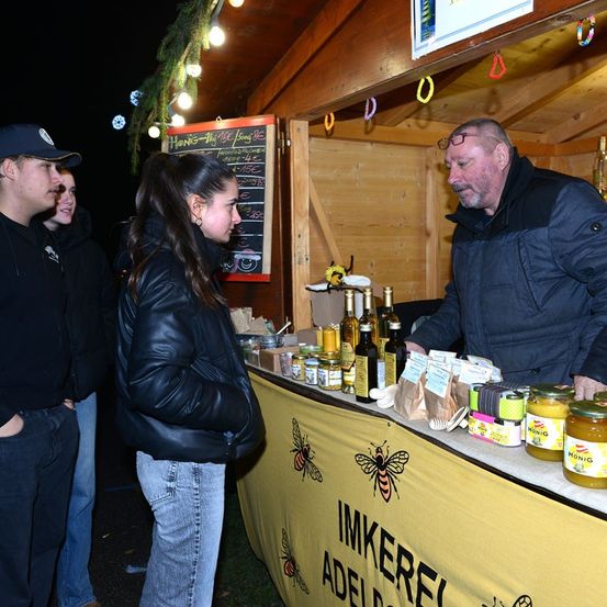 Ein Marktstand im Freien präsentiert Gläser mit Honig und anderen Produkten. Ein Mann und eine Frau unterhalten sich mit dem Verkäufer, der hinter einem Tisch mit einem gelben Banner steht.