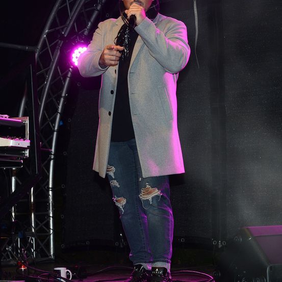 Ein Mann steht auf der Bühne unter einem Scheinwerfer und singt in ein Mikrofon. Er trägt einen grauen Mantel und zerrissene Jeans.