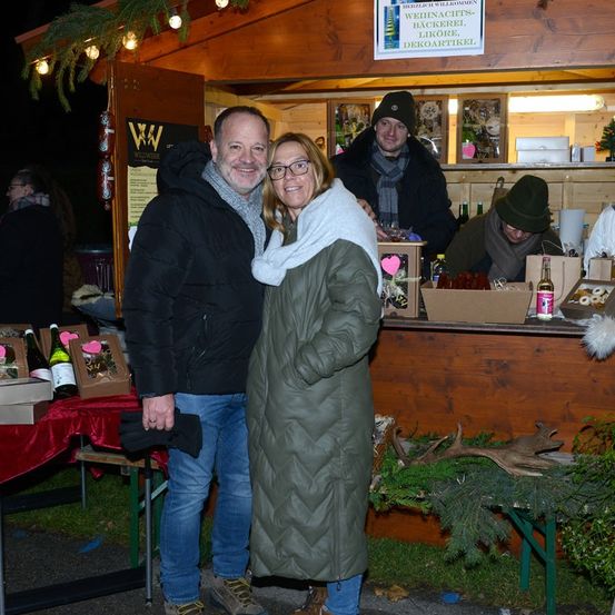 Ein Paar steht lächelnd vor einem hölzernen Laden mit Weihnachtsthema. Sie sind von verschiedenen Gegenständen umgeben, darunter Flaschen und Schachteln. Hinter ihnen sitzt ein Mann und ein anderer steht. Der Laden ist mit Weihnachtslichtern und Ornamenten geschmückt.